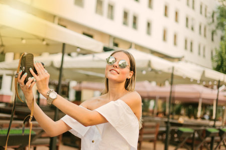 Cheerful fashionable woman in sunglasses takes pictures of herself on retro camera in urban environmentの写真素材