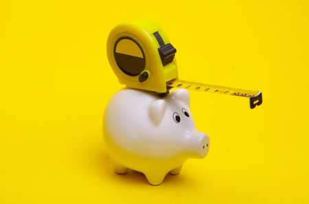 Ceramic white piggy bank with measuring tape on a yellow background. Conceptual photo