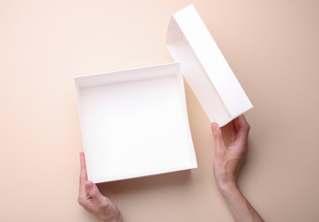 Female hands open white cardboard box on beige background. Mockup for design. Top viewの写真素材