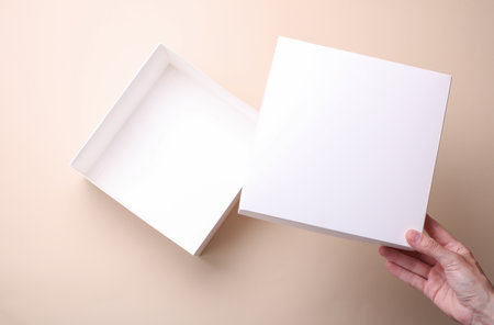 Female hands open white cardboard box on beige background. Mockup for design. Top view