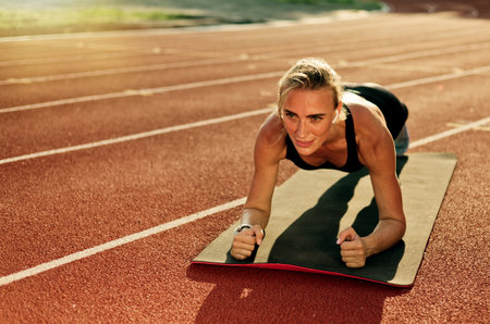 Attractive fit woman in sportswear doing plank exercise on stadium track with red coating on bright sunny dayの写真素材