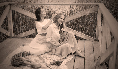Two cheerful young women have a picnic outdoors on a summer day. Two girlfriends in retro vintage style clothes spend time together on the pond pier.の写真素材