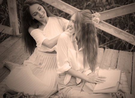 Two cheerful young women have a picnic outdoors on a summer day. Two girlfriends in retro vintage style clothes spend time together on the pond pier.の写真素材