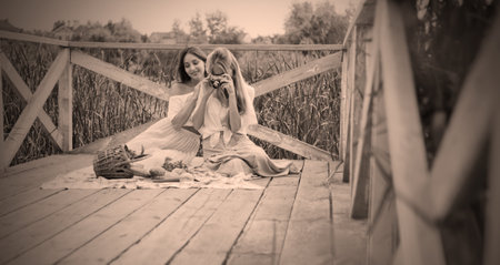 Two cheerful young women have a picnic outdoors on a summer day. Two girlfriends in retro vintage style clothes spend time together on the pond pier.の写真素材