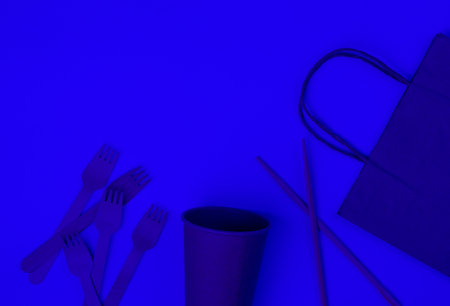 Disposable tableware of natural materials. Eco-friendly concept. Wooden forks, empty craft coffee cup, bag, chopsticks. Blue neon light. Copy space. Top viewの写真素材