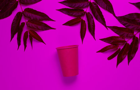 Environmentally friendly still life. Disposable craft cardboard coffee cup in pink neon light with green tropical leaves.の写真素材