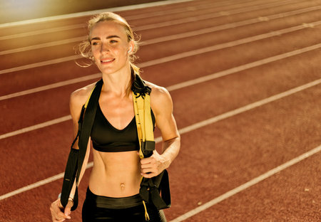 Young attractive woman athlete in sport clothes posing with fit straps on her neck at stadium track...