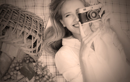 Retro style picnic. Young cheerful blonde woman in vintage clothes holding retro camera and taking photo while lies on picnic tablecloth outdoors. Top viewの写真素材