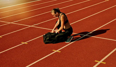 Young woman athlete in sportwear getting ready for workout and opening sports bag on the track of stadium at bright sunny dayの写真素材