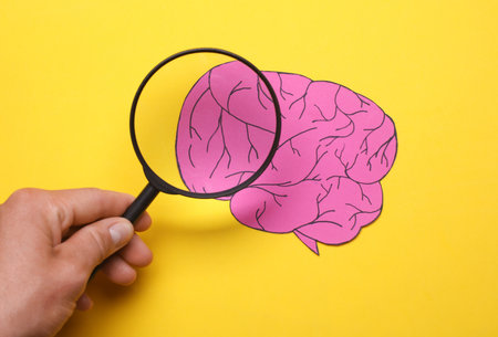 Hand with a magnifying glass examines paper cut brain on yellow background