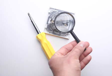 Hand with a magnifying glass examines Light switch with screwdriver. Repair
