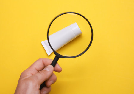 Hand holding magnifying glass and looking at cream tube with magnification, yellow backgroundの写真素材