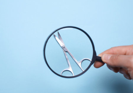 Hand with a magnifying glass examines manicure scissors for cleanliness on blue backgroundの写真素材