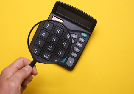 Hand holding magnifying glass and looking at calculator with magnification, yellow background