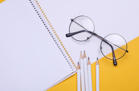 Sketchbook with pencils and eyeglasses on yellow background