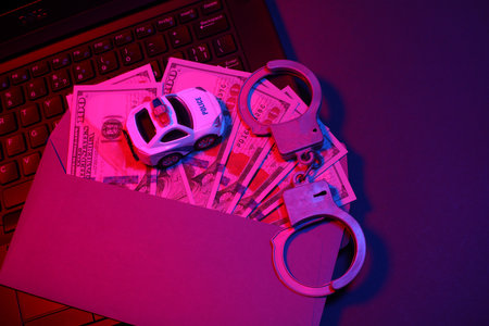 Laptop with handcuffs and dollar bills in envelope, toy police car in neon light. Cyber...