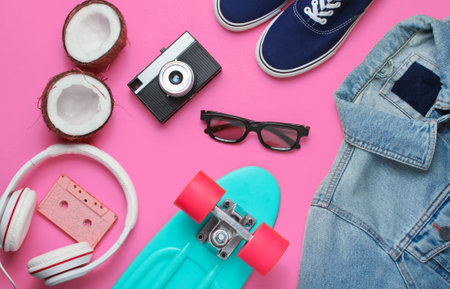 Hipster outfit. Skateboard with headphones, jeans jacket, audio cassette, retro camera, coconut and sneakers on pink background. Creative fashion minimalism. Minimal summer fun. 80s. Top viewの写真素材
