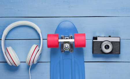 Hipster outfit. Skateboard, headphones, sneakers, retro camera on blue wooden background. Creative fashion minimalism. Minimal summer fun. Pop art. 80s. Copy space. Top viewの写真素材