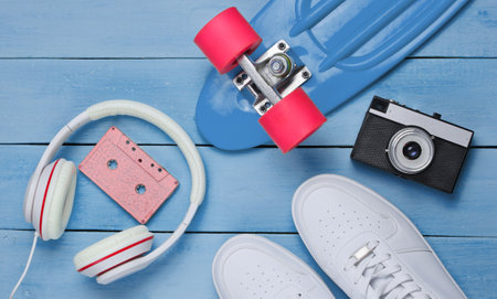 Hipster outfit. Skateboard, headphones, audio cassette, sneakers, retro camera on blue wooden background. Creative fashion minimalism. Minimal summer fun. Pop art. 80s. Copy space. Top viewの写真素材