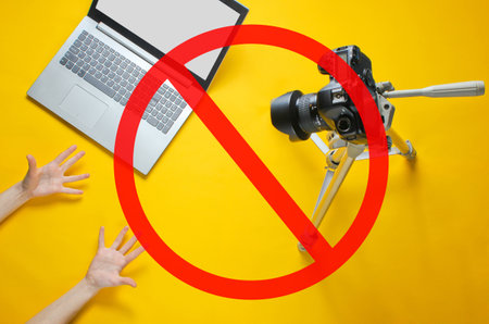 Online blogger concept. Emoticon reviewer. Women's hands show emotionally blog with camera on tripod, laptop on yellow background. Prohibition sign. Minimalism.の写真素材