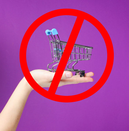 Mini toy metal shopping trolley on female hand on purple background with prohibition sign.の写真素材