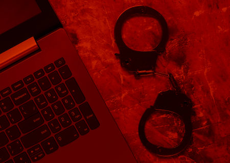 Cybercrime, online digital theft. Laptop with steel handcuffs in red light. Top viewの写真素材