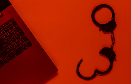Cybercrime, online digital theft. Laptop with steel handcuffs in red light. Top viewの写真素材