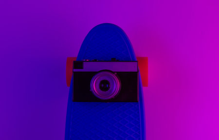 Plastic mini cruiser board and retro camera on background with blue pink neon gradient light. Top viewの写真素材
