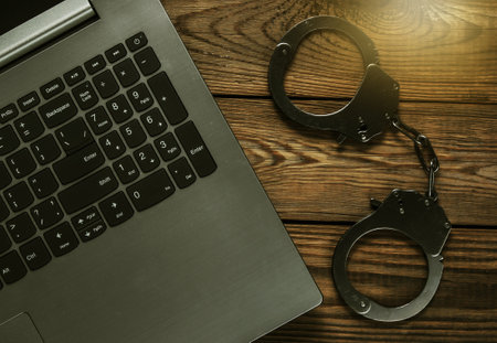 Cybercrime, online digital theft. Laptop with steel handcuffs on wooden table. Top viewの写真素材