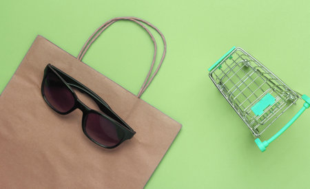 Minimalism shopping concept. Paper shopping bag, mini shopping cart and sunglasses on green background. Top viewの写真素材