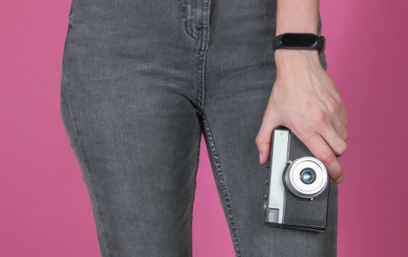 Woman in jeans holds a retro film camera on pink background. Crop photo