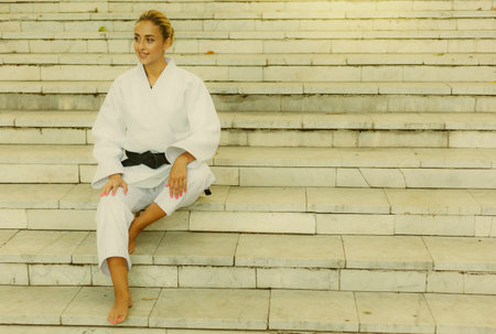 Young attractive woman in white kimono with black belt. Sport woman sitting on stairs outdoors. Martial artsの写真素材