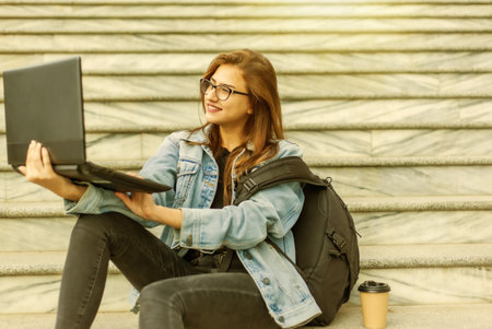 Young modern woman student in denim jacket and glasses sitting on the stairs with laptop. Watching video. Distance learning. Modern youth concept.の写真素材