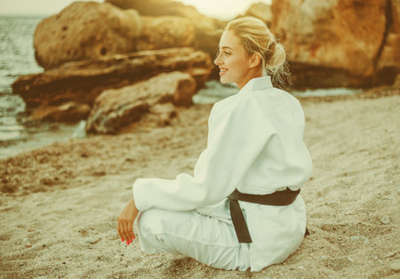 Young attractive karate woman in white kimono with black belt sitting on sand on wild beach with stonesの写真素材
