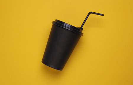 Black disposable cup for hot drinks with straw and lid on yellow backgroundの写真素材