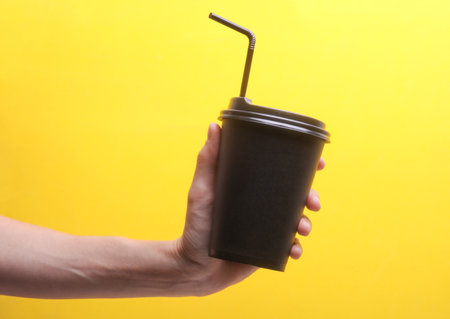 Hand holding Black disposable cup for hot drinks with straw and lid on yellow backgroundの写真素材