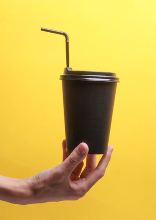 Hand holding Black disposable cup for hot drinks with straw and lid on yellow backgroundの写真素材