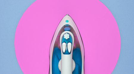 Modern iron for ironing on blue background with pink circle in the middle. Flat lay fashion still life. Top viewの写真素材