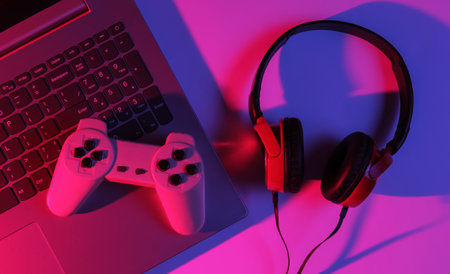 Gaming concept. Gaming equipment. Laptop with gamepad, headphones. Neon red-blue gradient light. Top...