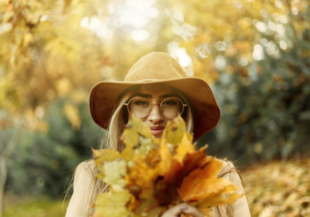 Young funny woman dressed in a coat and hat holds an armful of fallen leaves in the autumn park....
