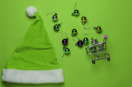 Christmas shopping. Santa hat, shopping cart, christmas balls on green background. Top view