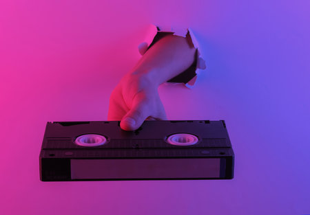 Hand holding video cassette through torn hole. Creative pop art pink blue neon color. Trendy...