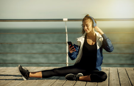 Relaxed fit woman in sportswear with headphones chooses music from a smartphone for training on the beach at bright sunny dayの写真素材