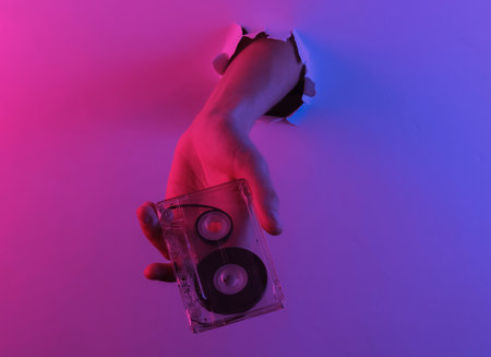 Hand holding audio cassette through torn hole. Creative pop art pink blue neon color. Trendy gradient illumination. Night lightの写真素材