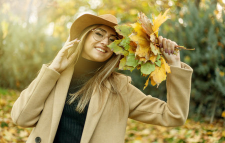Young funny woman dressed in a coat and hat holds an armful of fallen leaves in the autumn park....