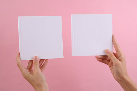Female hands holding square books with white blank cover for your design on pink background. Mockup