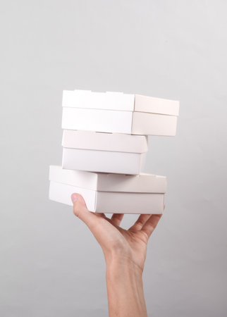 Female hand holding stack of white cardboard boxes on gray background. Food delivery, branding,...