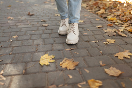 Female legs in light jeans and white leather boots. Woman walking in autumn park during daytime with fallen leavesの写真素材