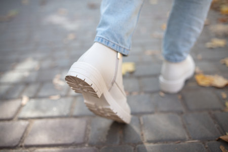 Female legs in light jeans and white leather boots. Woman walking in autumn park during daytime with...