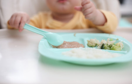 Cute little baby eats food from a plate at a tableの写真素材